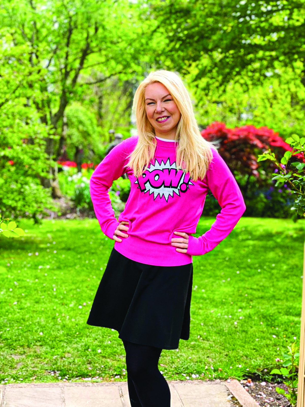 A portrait of Lauren Wright, standing in a garden, wearing a bright pink jumper with a comic book style 