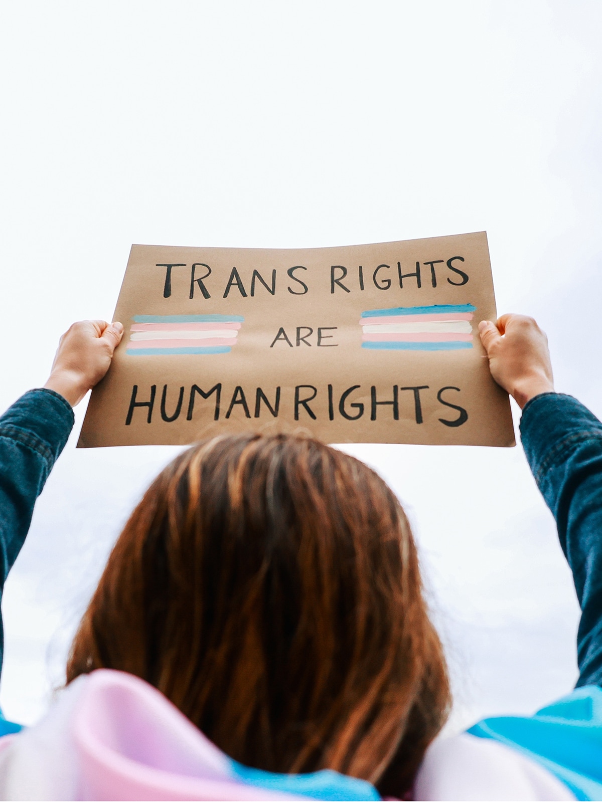 A person with long hair, wearing a trans rights flag, holds up a sign which reads 'Trans rights are human rights